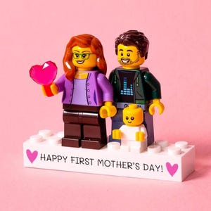 Op de afbeelding: Een Lego familie figuur met een moeder, vader en baby. De moeder houdt een roze hart vast en de basis leest "HAPPY FIRST MOTHER'S DAY!" in witte tekst. De figuren staan op een witte basis met roze harten.