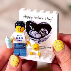 May include: A Lego-style figurine set with a white base and a sign that says "Happy Father's Day!" with a sonogram image. The set includes a "Best Dad Ever" figure holding a baby bottle and a small baby figure.