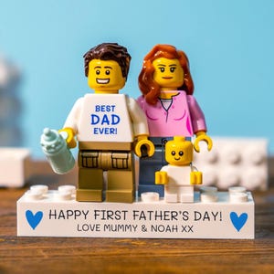 May include: A Lego family figurine set. The father figure wears a white shirt with "BEST DAD EVER!" printed on it and holds a baby bottle. The mother figure wears a pink shirt. The child figure is in white. The base reads "HAPPY FIRST FATHER'S DAY! LOVE MUMMY & NOAH XX".