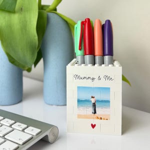 May include: A white brick pen holder with the text "Mummy & Me" and a photo of two people on a beach. The holder contains four colorful markers: green, red, orange, and purple. A red heart is below the photo.