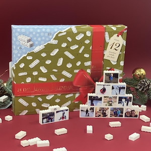 May include: A green and blue cardboard advent calendar with a red ribbon and bow. The calendar has 24 numbered doors, each with a small white Lego brick design. The calendar is labeled "24 Day Advent Calendar". There are also several small white Lego bricks scattered around the calendar.