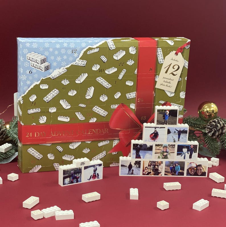 May include: A green and blue cardboard advent calendar with a red ribbon and bow. The calendar has 24 numbered doors, each with a small white Lego brick design. The calendar is labeled "24 Day Advent Calendar". There are also several small white Lego bricks scattered around the calendar.