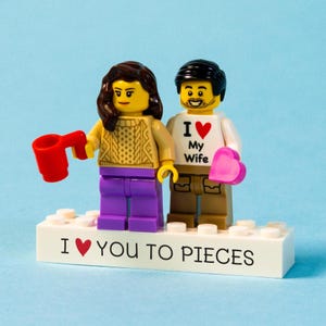 Può includere: Figurina di coppia Lego con una base bianca che recita "I ❤️ YOU TO PIECES". La figura femminile tiene in mano una tazza rossa, e la figura maschile tiene in mano un cuore rosa. La maglietta della figura maschile dice "I ❤️ My Wife."