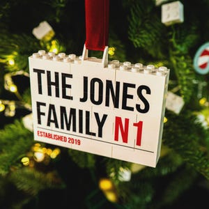 May include: A white rectangular ornament made of toy bricks, hanging from a red ribbon. The ornament reads "THE JONES FAMILY N1 ESTABLISHED 2019" in black and red lettering. The ornament is hanging on a Christmas tree.