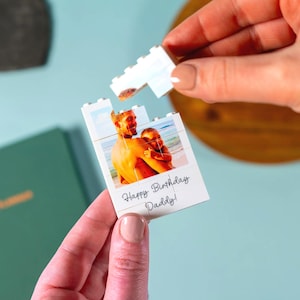 May include: A white puzzle piece with a photo of a man and a child, with the text "Happy Birthday Daddy!". The puzzle is being assembled by a person. The background is light blue.