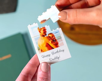 May include: A white puzzle piece with a photo of a man and a child, with the text "Happy Birthday Daddy!". The puzzle is being assembled by a person. The background is light blue.
