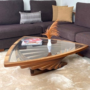 Puede incluir: Una mesa de centro triangular con tapa de cristal y base de madera hecha de piezas curvas apiladas. La mesa tiene un acabado marrón claro y está sobre una alfombra beige. Un jarrón con flores secas está sobre la mesa.