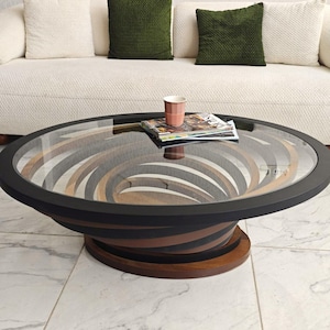 May include: A contemporary coffee table with a circular glass top. The table has a distinctive design with swirling wooden sections in brown and black. A magazine and a pink cup are on the table, which is in front of a white sofa with green and white cushions.