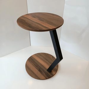 May include: A contemporary side table featuring a circular walnut top and base, linked by a black metal angled support. The table's design is minimalist, showcasing natural wood grain. The table's height is approximately 50 cm.