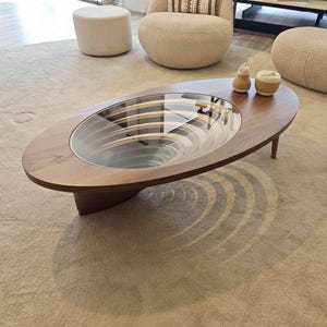 May include: A wooden oval coffee table with a glass top. The table has a unique design with a circular pattern that creates a swirling effect. The table is sitting on a beige rug.