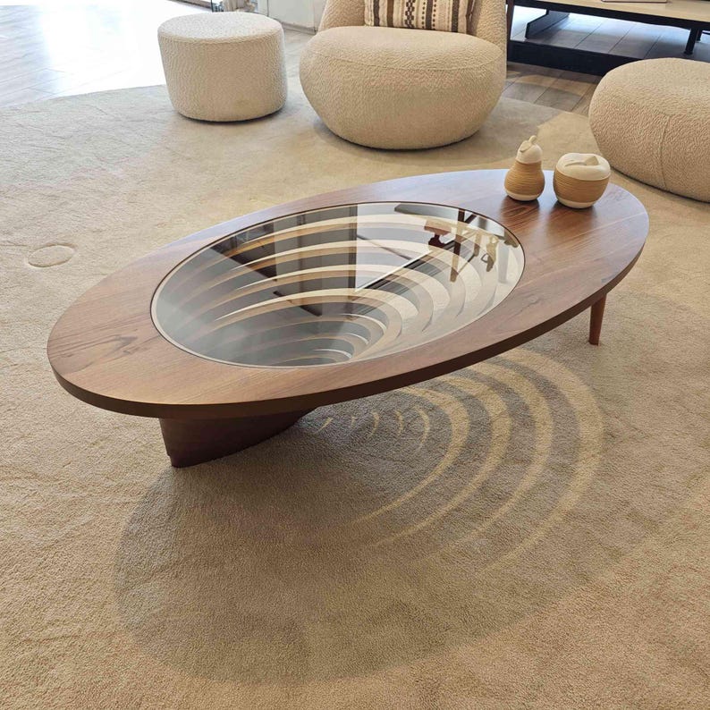 May include: A wooden oval coffee table with a glass top. The table has a unique design with a circular pattern that creates a swirling effect. The table is sitting on a beige rug.