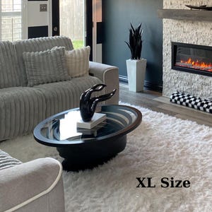 Peut inclure: Une table basse ovale noire avec un plateau en verre et une sculpture noire de mains. La table est pos&eacute;e sur un tapis blanc et duveteux. Un canap&eacute; gris avec des coussins est en arri&egrave;re-plan. Le texte "XL Size" est visible.