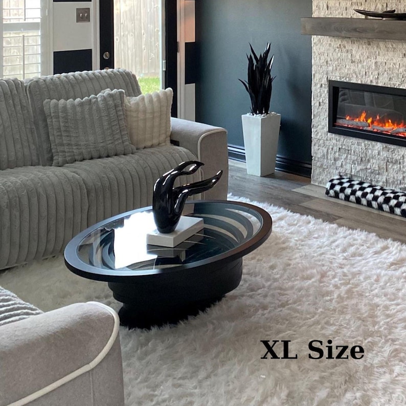 Peut inclure: Une table basse ovale noire avec un plateau en verre et une sculpture noire de mains. La table est pos&eacute;e sur un tapis blanc et duveteux. Un canap&eacute; gris avec des coussins est en arri&egrave;re-plan. Le texte "XL Size" est visible.