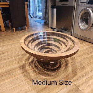 Peut inclure: Une table basse ronde de taille moyenne avec un design en spirale unique. Elle est dot&eacute;e d'un plateau en verre transparent et d'une finition en bois brun clair. La base pr&eacute;sente un design concentrique en couches. Le texte "Medium Size" est visible.