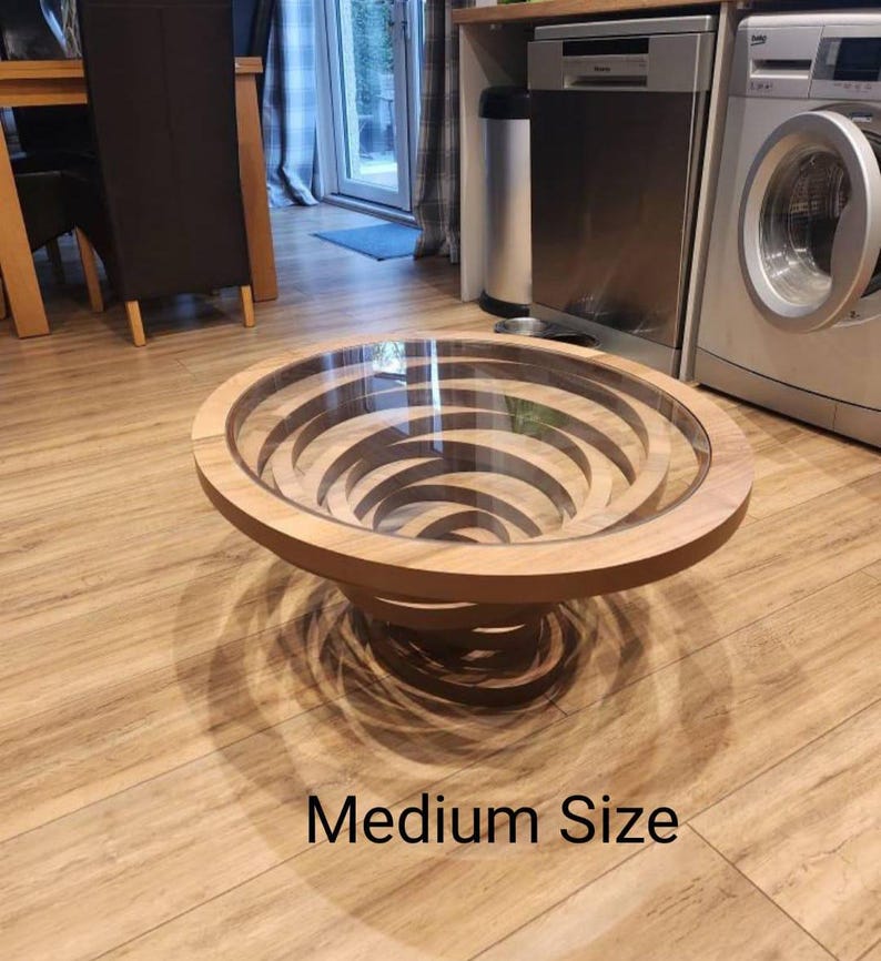 Peut inclure: Une table basse ronde de taille moyenne avec un design en spirale unique. Elle est dot&eacute;e d'un plateau en verre transparent et d'une finition en bois brun clair. La base pr&eacute;sente un design concentrique en couches. Le texte "Medium Size" est visible.