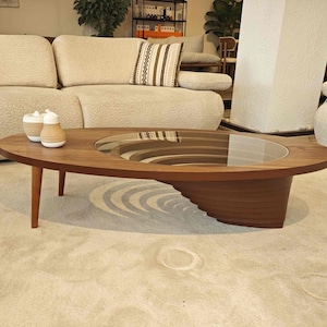 May include: A modern oval-shaped coffee table with a glass top and a unique layered wooden base. The table is made of dark wood and has a natural finish. The table is sitting on a light beige rug.