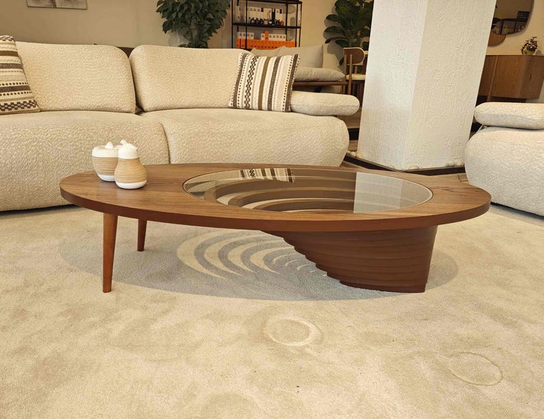 May include: A modern oval-shaped coffee table with a glass top and a unique layered wooden base. The table is made of dark wood and has a natural finish. The table is sitting on a light beige rug.