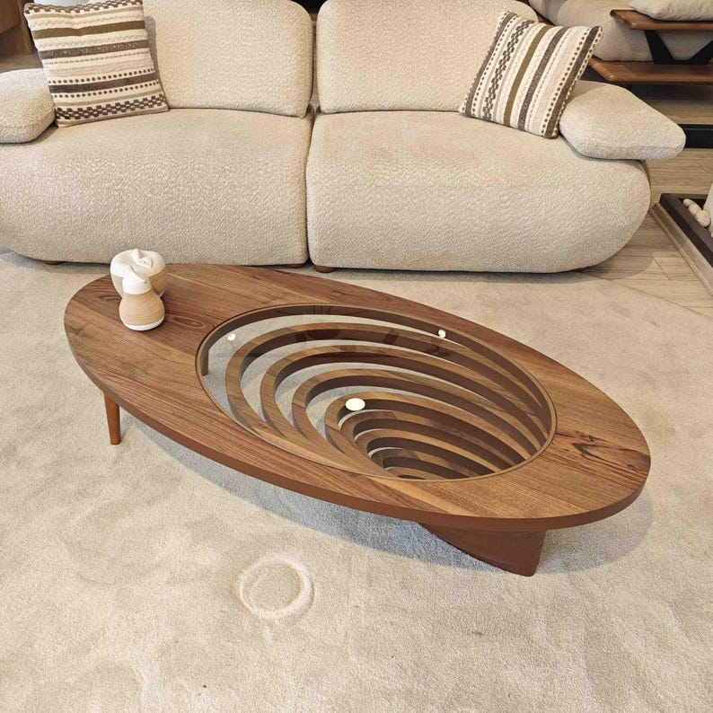 May include: Oval-shaped wooden coffee table with a unique design featuring a glass top and a series of concentric rings carved into the wood, creating a swirling effect.