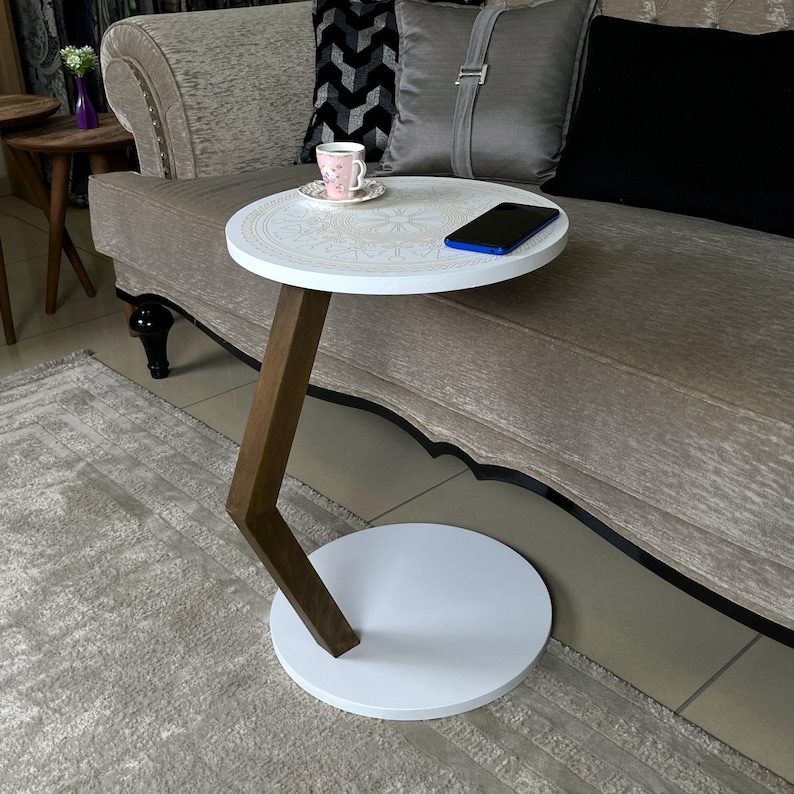 May include: A white side table with a round top and base, connected by a dark brown, angled support. The tabletop features a decorative floral design. A smartphone and a teacup and saucer are on the table.