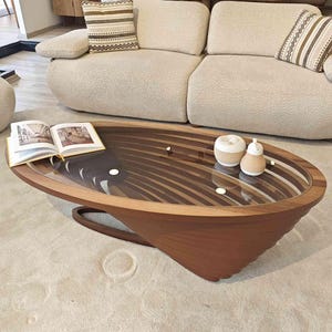 May include: A wooden oval coffee table with a glass top and a unique, layered design. The table has a dark brown finish and is sitting on a light beige rug.