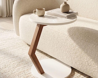 Puede incluir: Una mesa auxiliar en forma de C con una superficie redonda blanca que presenta un patrón de mandala grabado, una pata de soporte angular de madera oscura y una base circular blanca. La mesa sostiene una taza de cerámica beige, un jarrón y dos libros.