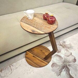May include: A round wooden side table with a decorative carved top, supported by a single angled leg and a circular base. The table holds a white teacup and two red and gold decorative pomegranates. The table is near a beige sofa.
