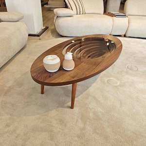 May include: A wooden oval coffee table with a unique spiral design in the center. The table has a glass top and is sitting on a beige carpet. There are two decorative objects on the table, a white ceramic apple and a white ceramic pear.