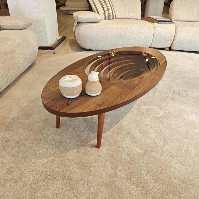 May include: A wooden oval coffee table with a unique spiral design in the center. The table has a glass top and is sitting on a beige carpet. There are two decorative objects on the table, a white ceramic apple and a white ceramic pear.