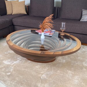 Peut inclure: Table basse ovale avec un plateau en verre transparent et une base en bois au design unique. La base pr&eacute;sente un motif en spirale dans les tons bruns. Un petit vase blanc avec des fleurs s&eacute;ch&eacute;es et un magazine sont sur la table.