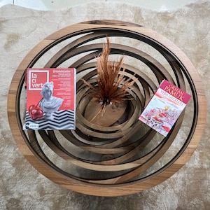 Peut inclure: Une table basse en bois avec un design en spirale. La table pr&eacute;sente une finition en bois fonc&eacute; et contient un magazine avec une couverture rouge et un livre. Un vase avec des plantes s&eacute;ch&eacute;es est au centre.