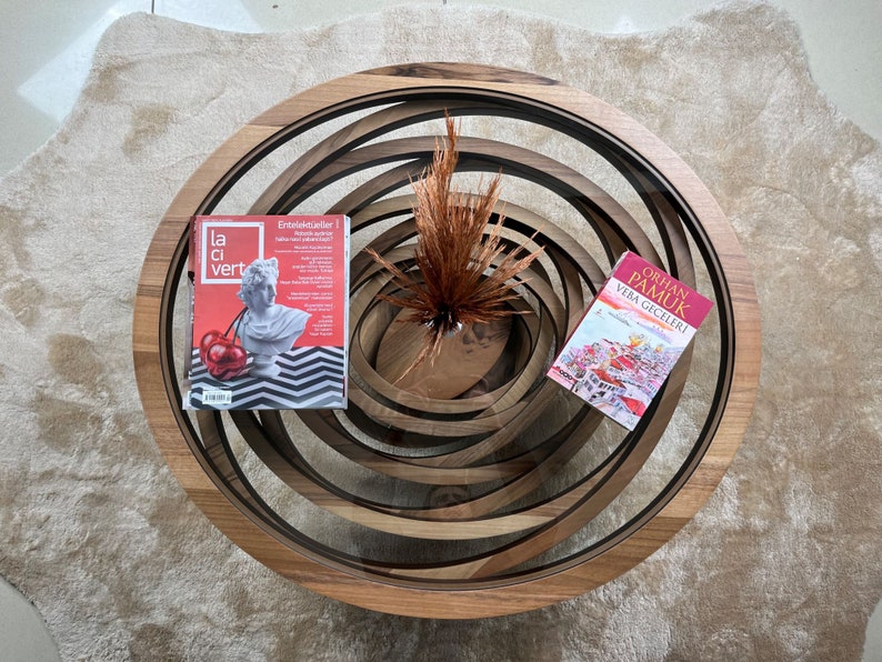 Peut inclure: Une table basse en bois avec un design en spirale. La table pr&eacute;sente une finition en bois fonc&eacute; et contient un magazine avec une couverture rouge et un livre. Un vase avec des plantes s&eacute;ch&eacute;es est au centre.