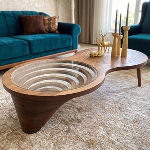 May include: A wooden coffee table with a unique figure-eight shape, featuring a circular glass top insert over a recessed, concentric ring design. The other side of the table surface displays a carved, circular pattern with three tall candle holders.