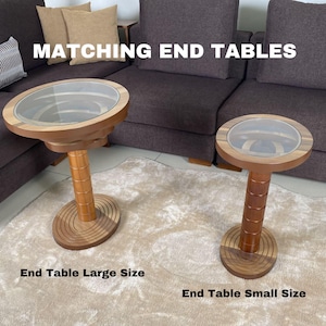 May include: Matching wooden end tables with glass tops. The larger table is on the left and the smaller table is on the right. Both tables have a circular top and a cylindrical base.