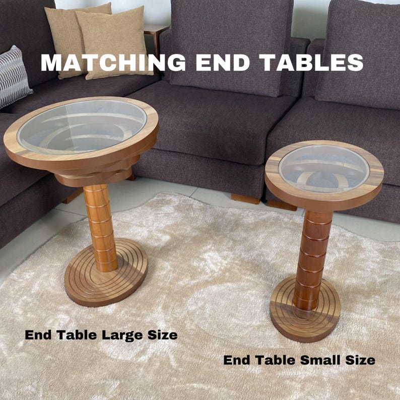 May include: Matching wooden end tables with glass tops. The larger table is on the left and the smaller table is on the right. Both tables have a circular top and a cylindrical base.