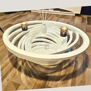 May include: A round coffee table featuring a clear glass top and a white base constructed from multiple concentric, spiraling rings. Two dark, cylindrical candle holders with light-emitting patterns and a white reed diffuser sit on top.
