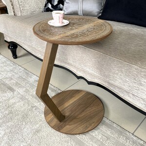 May include: A wooden side table with a round top and base, connected by a slanted, angular support. The tabletop features an etched, circular design. A cup and saucer sit on the table. The table is approximately 61 cm tall.