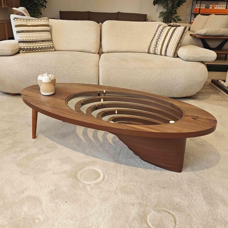 May include: A dark wood oval coffee table with a unique design featuring a glass top and concentric circles cut into the wood. The table has a modern, minimalist style and would be a great addition to any living room.