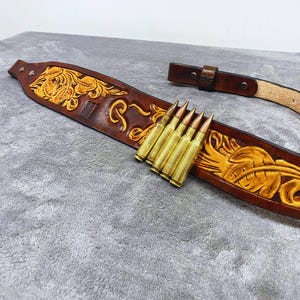 Op de afbeelding: Een bruine leren riem met ingewikkelde bloemmotieven en een rij messing hulzen. De riem heeft een gesp en een lus. De bewerking is geaccentueerd met een lichtere bruine kleur.