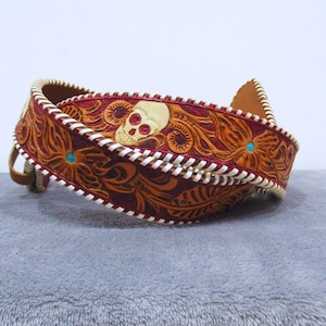 May include: A decorative leather belt with a carved floral design in shades of brown and burgundy. The belt features a skull and turquoise accents. The edges are stitched with white thread.