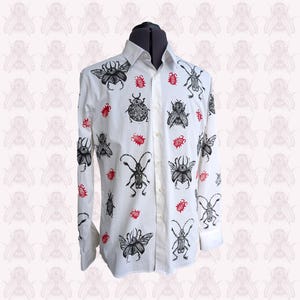 Op de afbeelding: Wit overhemd met zwarte insectenpatroon en rode accenten. Het shirt met lange mouwen heeft verschillende insectenillustraties, waaronder kevers en vliegen. Het shirt wordt gepresenteerd op een zwarte mannequin.