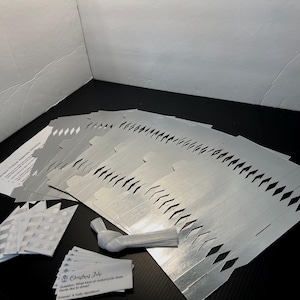 May include: A set of silver metallic DIY Christmas cracker templates with die-cut diamond patterns, white paper ties, a sheet of adhesive dots, and small printed joke cards featuring the text Christmas Joke with a question and answer.