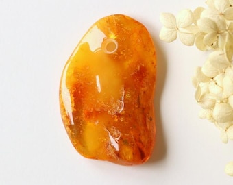 May include: A polished amber pendant with a hole for stringing. The pendant is a warm, translucent orange color with internal variations. Dried white flowers are in the background.