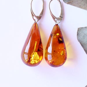 Op de afbeelding: Een paar amberkleurige druppelvormige oorbellen met zilverkleurige oorhaaksluitingen. De oorbellen hebben een warme, doorschijnende oranje tint met zichtbare insluitsels. Op een witte achtergrond.