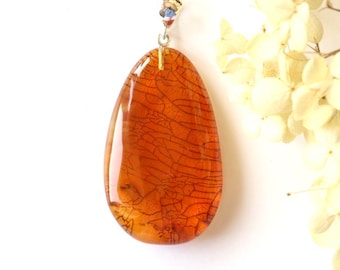 May include: A teardrop-shaped pendant with a warm orange hue and intricate dark vein-like patterns. The pendant hangs from a silver chain with a beaded accent. The background is white with a sprig of dried flowers.
