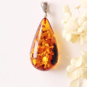 May include: A teardrop-shaped amber pendant with a silver bail. The amber is a translucent, golden-orange colour with dark inclusions. The pendant hangs against a white background, with dried white flowers in the background.