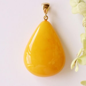 May include: A smooth, teardrop-shaped pendant with a vibrant, solid yellow, opaque finish. The pendant features a polished surface and is attached to a small, metallic gold-toned bail at the top, designed for suspension on a chain or cord.
