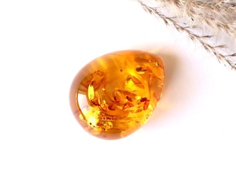 May include: A teardrop-shaped amber pendant with a golden yellow hue and inclusions of plant matter. The pendant is smooth and polished.