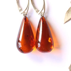 May include: A pair of drop earrings featuring polished, teardrop-shaped amber stones in a deep, translucent reddish-brown hue. The stones are suspended from silver-toned metal lever-back hooks, showcasing natural inclusions within the resin material.