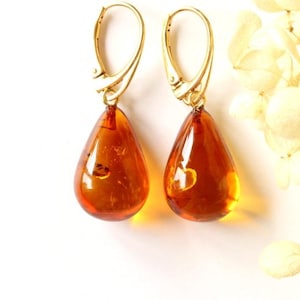 May include: A pair of amber teardrop earrings with gold-coloured hooks. The amber stones are a translucent, warm brown colour, with small inclusions visible within. The earrings are displayed against a white background.