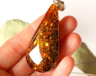 May include: A teardrop-shaped amber pendant with a silver bail. The amber is a deep orange color with flecks of gold and black throughout.